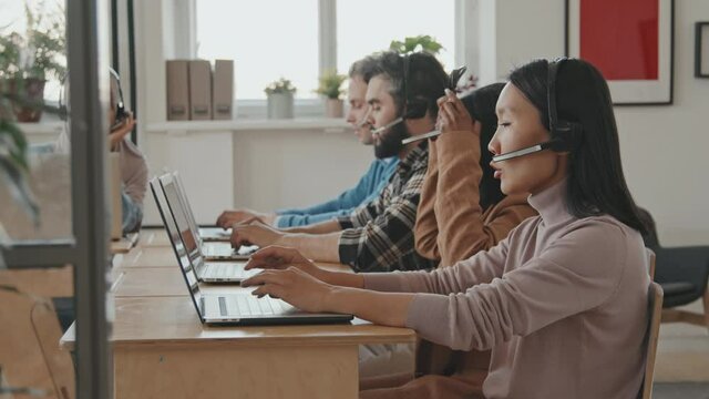 Tracking shot of diverse group of call center workers in headsets sitting at desks and talking to clients