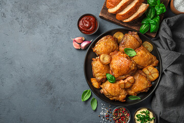 Dinner with fried chicken thighs with onions, garlic and fresh basil on a dark background with sauces, spices and bread.