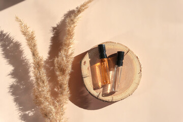 Two glass perfume samples with transparent brown liquid on wooden tray lying on beige background with pampas grass. Luxury and natural cosmetics presentation. Testers on woodcut in sunlight. Top view