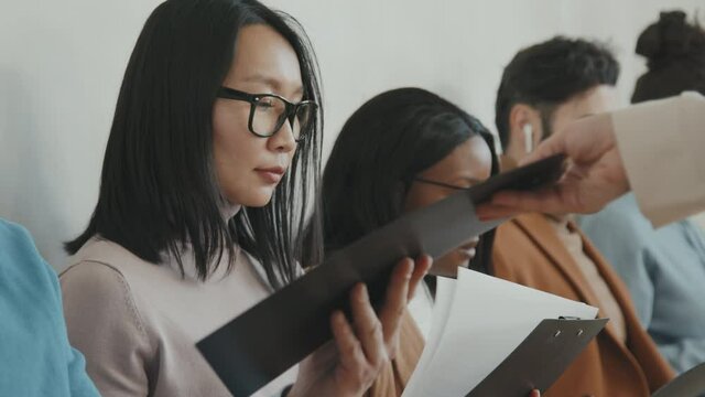Tracking medium shot of unrecognizable HR manager giving application forms to diverse group of people sitting in queue for job interview