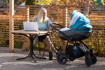 a young woman with  ​baby carriage working or studying on her laptop in outdoor cafe. remote work, study during maternity leave
