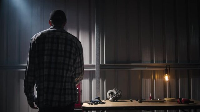 A tall man walking into an industrial garage with a light shining down, opening a red toolbox and taking out tools. On the workbench in the background you can see a turbocharger, gloves, hammer, work 