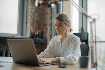 Young woman with laptop and phone at work table in modern office in the afternoon