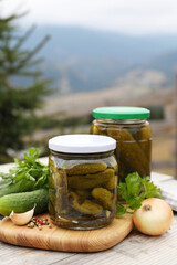 Jars of delicious pickled cucumbers and ingredients on wooden table in mountains, space for text
