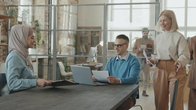 Tracking shot of male executive and female HR manager conducting job interview with cheerful Muslim woman in hijab. They are sitting down at table in office and having conversation