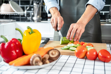 Asian man cooking wearing apron in the kitchen and using knife to slice cucumber and tomato on chopping board