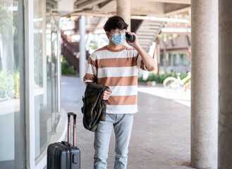 Tourist man in medical face mask is talking with friends on smartphone while standing with luggage for vacation