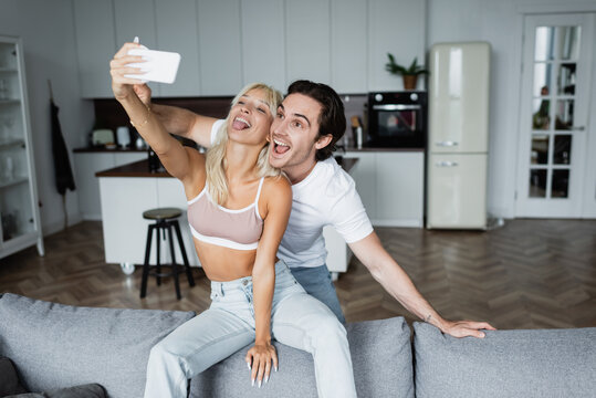 happy woman sticking out tongue while taking selfie with boyfriend in living room