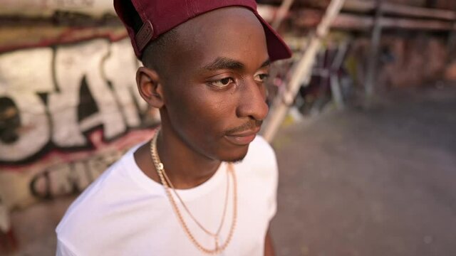 A young guy in a cap on the street in the ghetto posing against the background of a wall with graffiti in an alley. Dangerous area, disadvantaged area, suburb