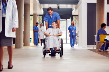 Fototapeta premium Male Nurse Wearing Scrubs Pushing Female Patient In Wheelchair Through Hospital Building