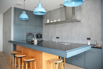 A modern kitchen design with gray walls, glass and gray kitchen cabinets, wooden stools, a black kitchen island, and blue lamps above. A kitchen interior with a built-in oven and coffee machine.