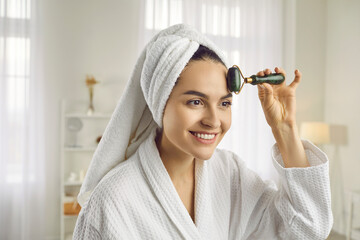 Obraz na płótnie Canvas Happy young woman in bathrobe and bath towel massaging face with trendy smooth jade facial roller to stimulate lymphatic system, relax muscles, reduce puffiness, increase blood flow for glowing skin