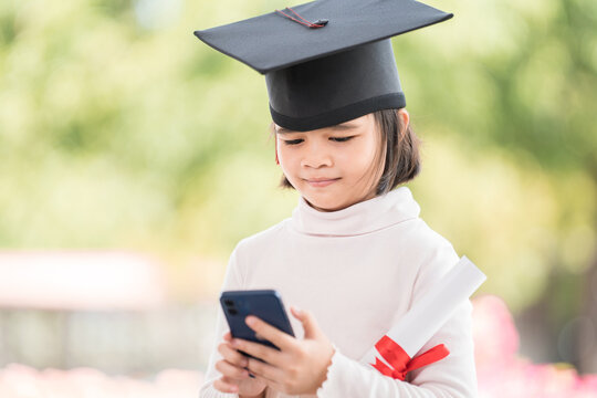 Happy Asian female kid graduate with graduation cap and rolled graduation certificate holding a smartphone to call and chat with friends and family. Education Concept Stock Photo