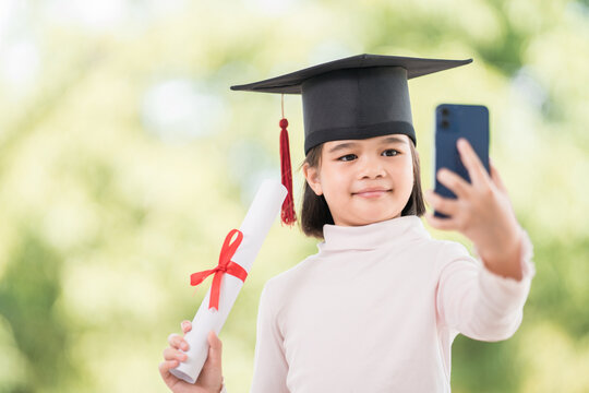 Happy Asian female kid graduate with graduation cap and rolled graduation certificate holding a smartphone to call and chat with friends and family. Education Concept Stock Photo