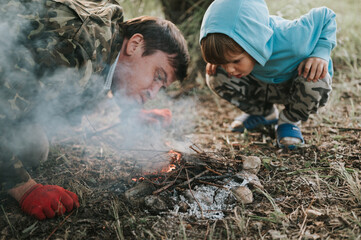 little happy five year old kid boy and his father blowing and kindles a campfire in the village or...