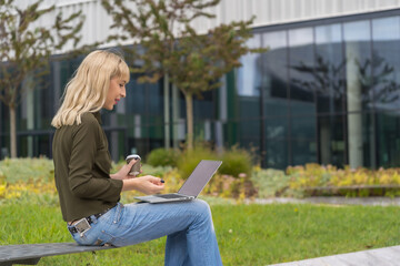 Caucasian blonde girl entrepreneur in business park conducting a video conference with a take away coffee. Modern polygon among nature