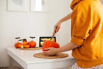 Woman in yellow hoodie making jack-o-lantern by carving a pumpkin on the table while watching a tutorial on tablet