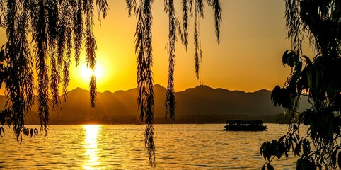 Sunset landscape and scenery at Hangzhou West Lake, sunlight shining through falling willow tree leaves as tour boat crosses the lake passing mountains © Adam