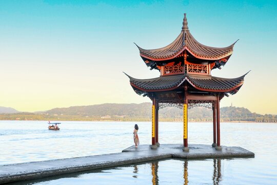 Beautiful sunrise at Hangzhou West Lake, elegant woman in dress stands by the Jixian Pavilion watching rowing boat in the vast lake