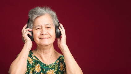 Cheerful elderly Asian woman with short gray hair wearing wireless headphones to listen to a favorite song with a smile while standing on a red background. Aged people and relaxation concept