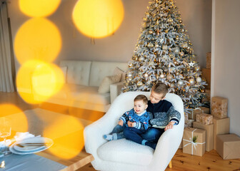 Loving brothers sittings near the Christmas tree with a lot of gifts. Happy New Year. Christmas morning in bright living room. Stylish fashion little boys wearing jackets. bokeh in the foreground
