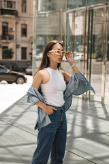 Fototapeta premium Half-length image of serious young woman standing in front of panoramic building. Brunette with glasses confidently looks in front of with her hand in her pocket. Lifestyle concept