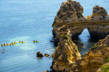 kayaks pass under a rocky arch on sandy beach Praia do Camilo in Lagos, Algarve, Portugal