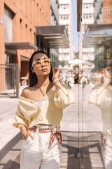  Sophisticated asian woman stands relaxed basking on sun in fresh air. calm girl in sunglasses is wearing light jacket and white pants with fabric belt. Harmony and tranquility concept