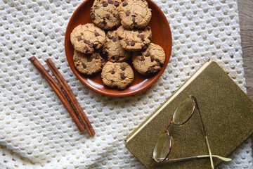 Plate of chocolate chip cookies, knitted blanket, book, reading glasses and cinnamon sticks. Hygge at home. Flat lay.