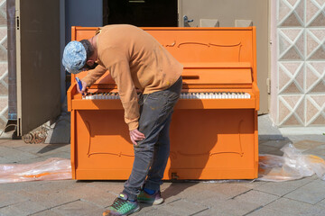 A man in a cap standing over an orange colored piano and fixing it