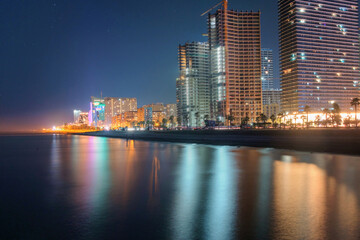 An illuminated night beach with modern architecture lights reflecting on the sea surface, view from the sea