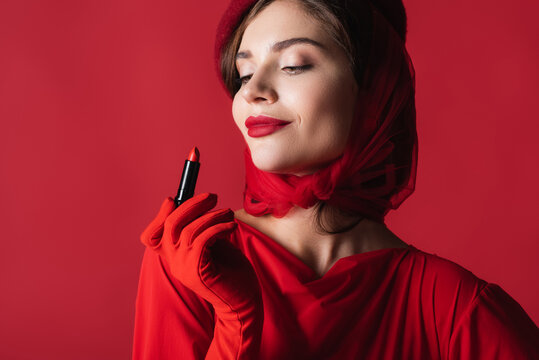 cheerful woman in glove, headscarf and beret looking at lipstick isolated on red.