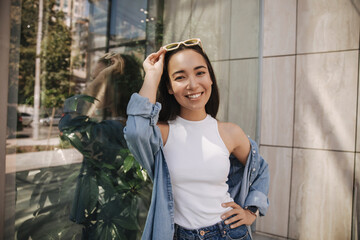 Close-up of young beautiful woman smiling at camera rejoicing walking through city streets on sunny summer day. Asian woman with loose long hair is dressed in white T-shirt, shirt and jeans.