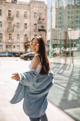 Fototapeta premium Full-length image of feminine asian on blurred background of city. Long-haired brunette in blouse and shirt top, sunglasses on her face. Good mood, fashion trends