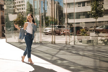 Full-length graceful asian lady walks with flying gait along road against backdrop of glass house. Brunette is talking on phone with her mouth parted. She is wearing light everyday clothes.