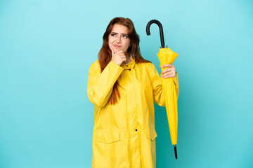 Redhead woman holding an umbrella isolated on blue background having doubts
