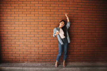 Fototapeta na wymiar In full growth happy young asian girl in glasses posing raising her hand up on brick background. She is wearing loose jeans, white T-shirt, blue shirt and sandals. Summer playful mood concept