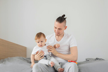 Smiling father sitting on bed holding his one year old son. White background.