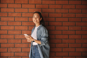 Young beautiful brunette in denim jacket near wall smiles, uses modern mobile phone. Long-haired asian tries to compose good message. Cell phone use concept