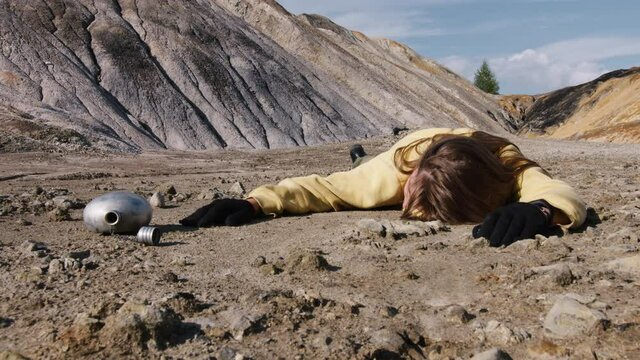 Girl exhausted from thirst lies in rocky wasteland with empty metal flask. Becoming weak hungry woman sprawled on ground. Drunk person is on sandy surface. Helplessness, early death in stone badlands