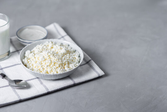Homemade cottage cheese with glass of milk, spoon in bowl on light background. Healthy dairy products, baking ingredients. Source of calcium for breakfast, diet food. Copy space for text