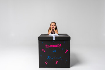 little cute emotional girl in a colored dress with blond hair leaned on a festive black box with a bow on a white background. She is thoughtful, smiling. Birthday, celebration, anticipation.