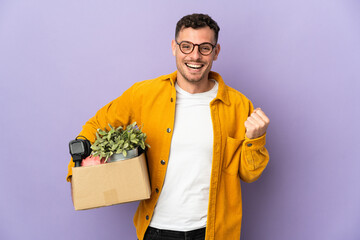 Young caucasian man making a move while picking up a box full of things isolated on purple...