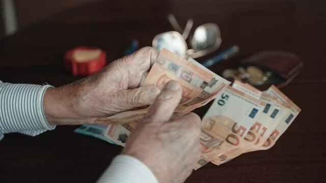 Old male pensioner sitting at the table, holding stack of paper currency and counting 50 euro banknotes. Senior man calculating his meager monthly pension payment, low financial income and pension