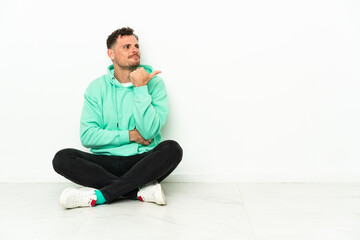 Fototapeta premium Young handsome caucasian man sitting on the floor pointing to the side to present a product