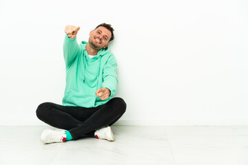 Obraz premium Young handsome caucasian man sitting on the floor points finger at you while smiling