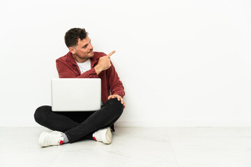 Young handsome caucasian man sit-in on the floor with laptop pointing back with the index finger