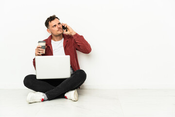 Young handsome caucasian man sit-in on the floor with laptop holding coffee to take away and a mobile