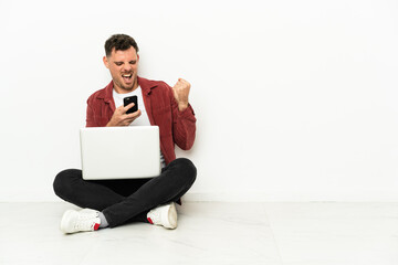 Young handsome caucasian man sit-in on the floor with laptop with phone in victory position