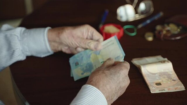 Poverty and low standard of living concept, old pensioner counting meager pension payment sitting alone in living room, senior man in retirement counting 20 euro paper currency, putting on the desk